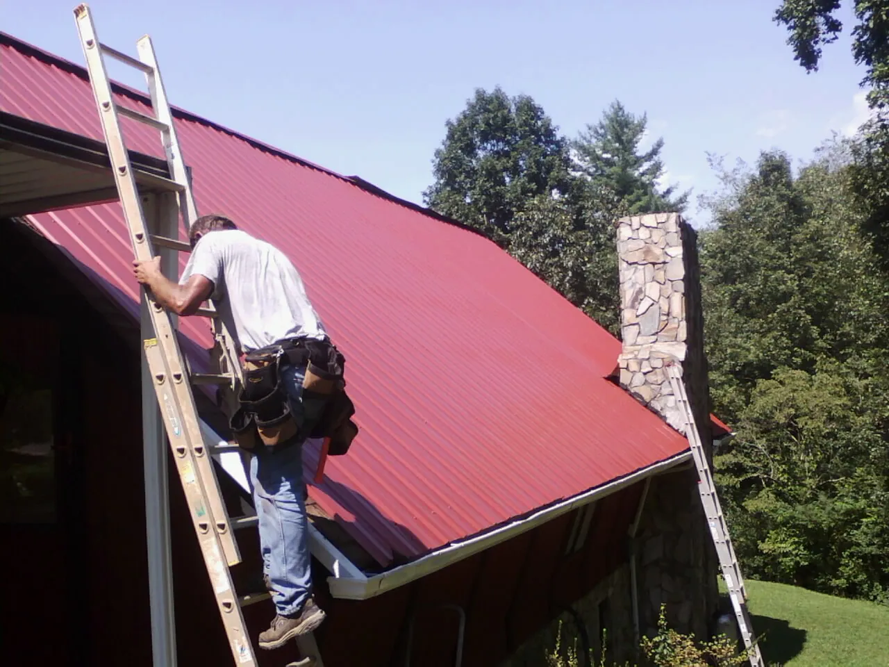 Roofer on a ladder beside a red metal roof and stone chimney — Soffit & Fascia Repair by Roofing Brothers in Federalsburg